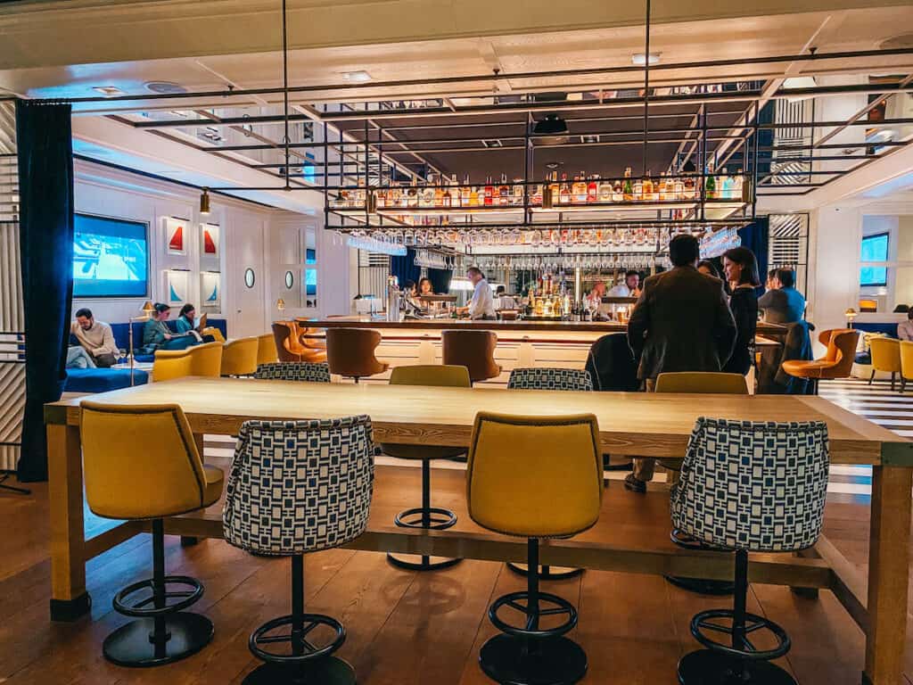 A hotel bar with stools and a bar in the middle, on the side you can see some tables