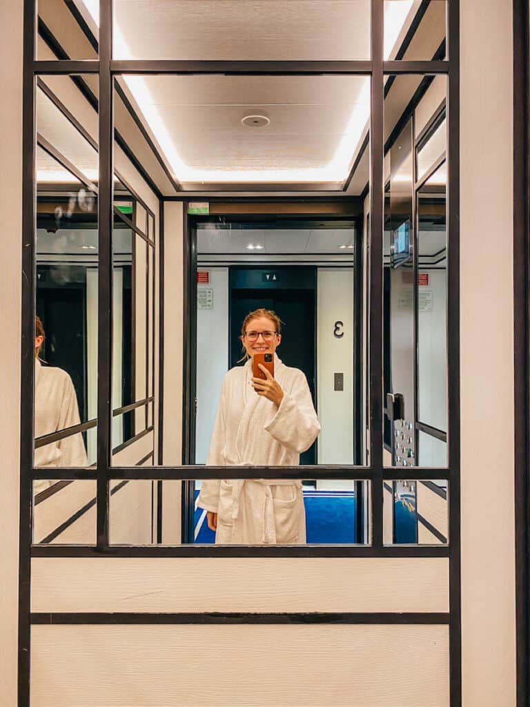 Tina taking a mirror selfie inside an elevator, she is just wearing a bathrobe