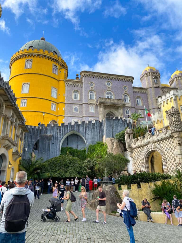 Colorful exterior of Pena Palace in Sintra, Portugal, featuring a vivid yellow domed tower and lavender-tiled facade with ornate details. Tourists walk and gather in the cobblestone courtyard below, surrounded by lush greenery and whimsical architectural elements under a bright blue sky.