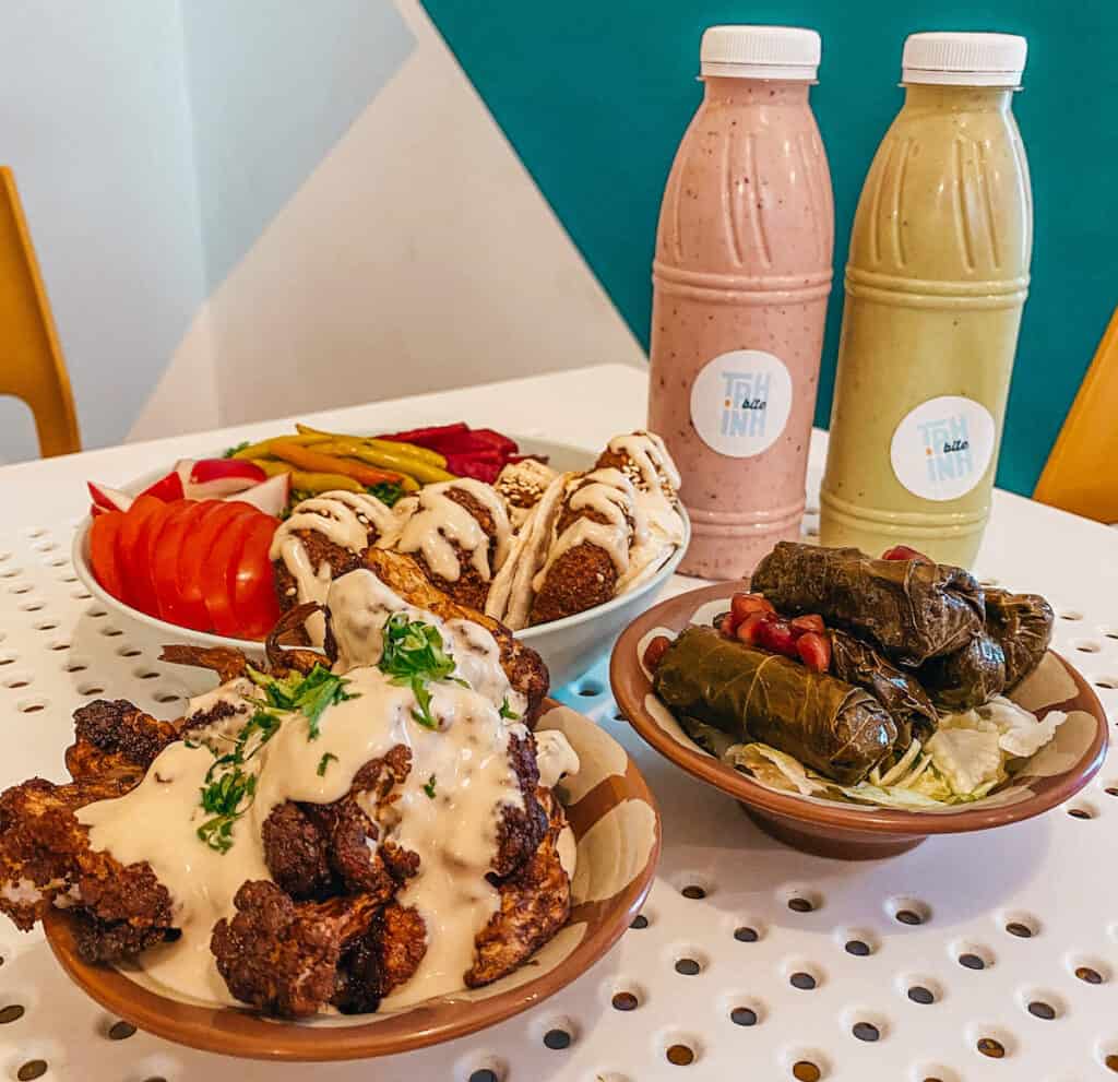 An assortment of vegan dishes including falafel wraps, fried cauliflower with sauce, stuffed grape leaves, and colorful pickled vegetables, flanked by two bottles of smoothies, presented on a white table with blue accents.