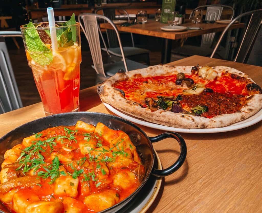A vibrant meal with a bowl of gnocchi in tomato sauce garnished with parsley, a wood-fired vegetable pizza, and a tall glass of iced tea with mint, served on a dark wooden table.