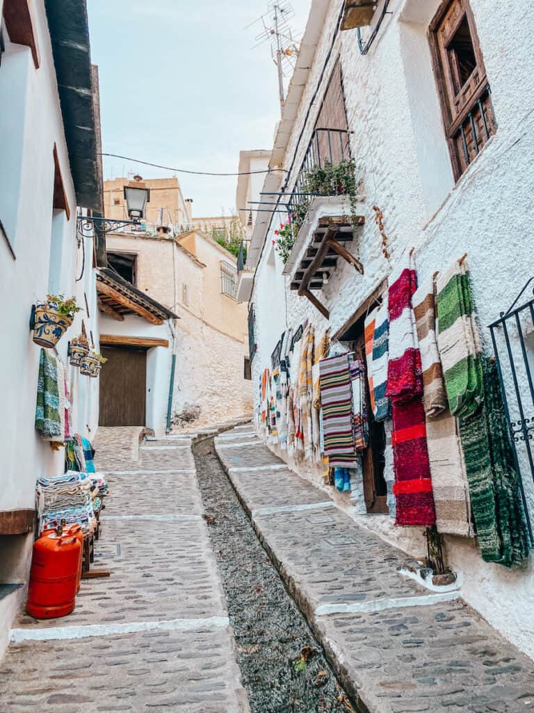 Charming narrow alley in a traditional white village in the Sierra Nevada, with whitewashed walls adorned with colorful textiles, flowers on balconies, and cobblestone pavement, reflecting the authentic cultural experience for a '10 days in Southern Spain' trip.