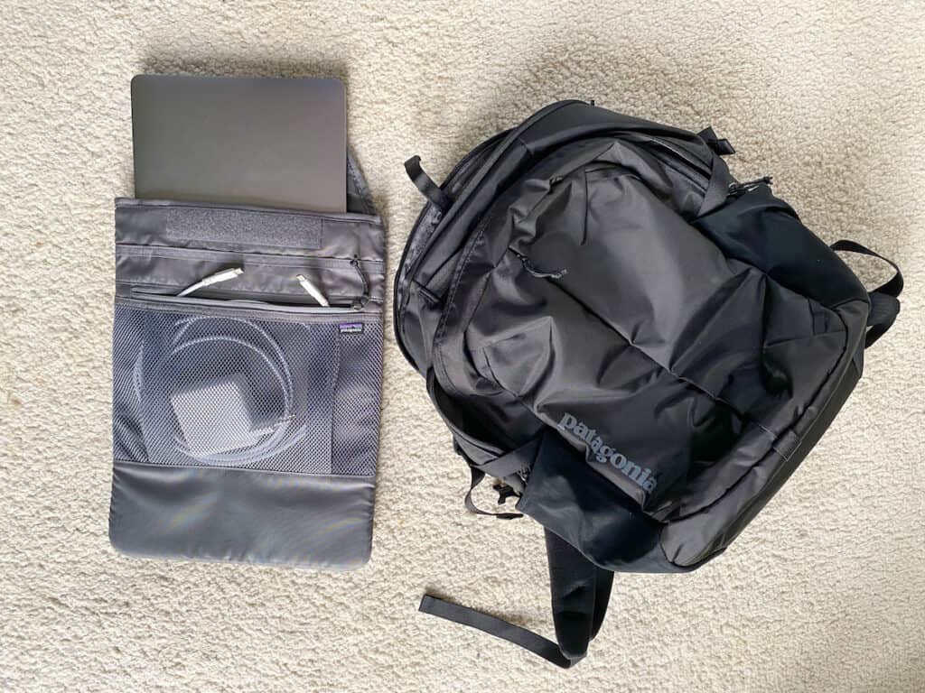 A Patagonia Refugio 26L backpack lying flat on a carpeted floor next to a grey laptop, showcasing its sleek design and spacious front compartment.