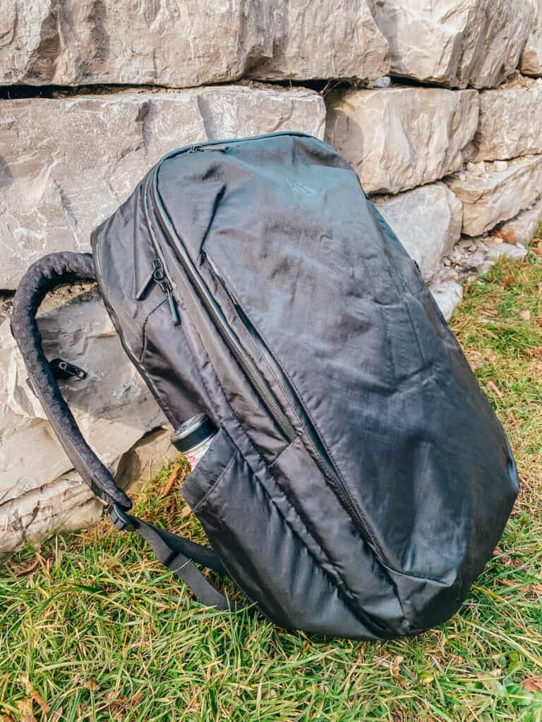 A Tortuga 30L travel backpack rests against a rugged stone wall on grassy terrain, with a water bottle peeking out from the side pocket, capturing the essence of outdoor adventures and the backpack's sturdy design.