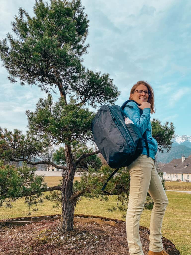 Tina standing beside a pine tree, with the Tortuga 30L backpack slung over one shoulder, ready for a hike in the scenic outdoors.