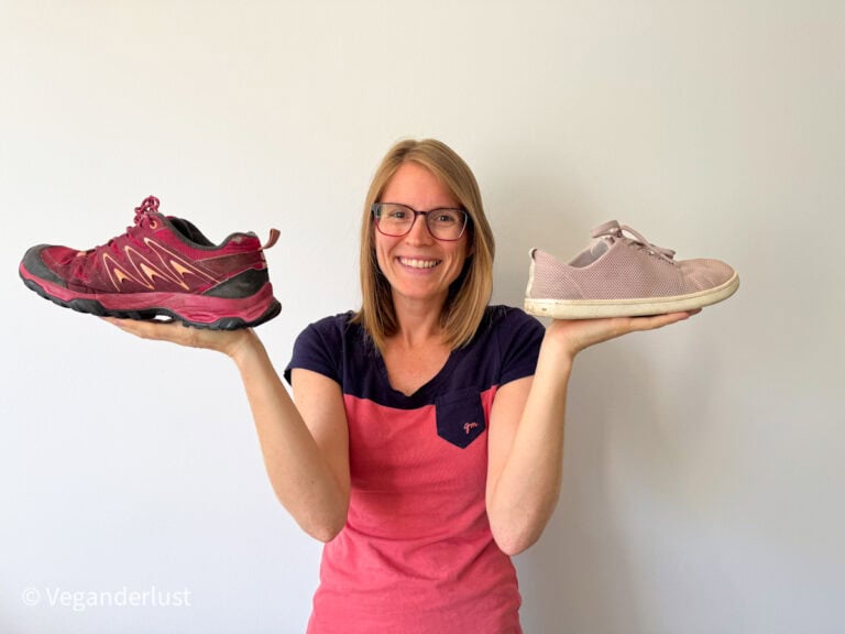 Smiling woman in a color-blocked pink and navy shirt holds a maroon trail shoe in one hand and a light pink casual sneaker in the other, comparing the two. She stands against a plain white wall, suggesting a choice between rugged outdoor footwear and lightweight travel shoes.