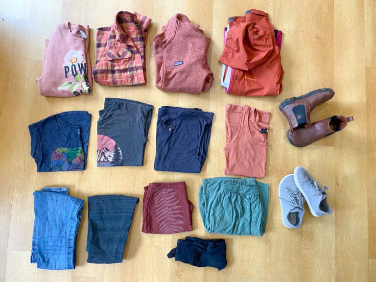 Assorted spring travel clothing laid out on a wooden floor, featuring a pink 'POWER' sweatshirt, plaid shirt, fleece pullover, and a selection of t-shirts, shorts, jeans, and footwear, all adhering to a minimalist packing list for Europe in spring.