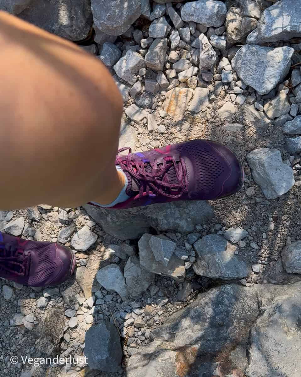 Close-up of a personโs legs and feet standing on a rocky hiking trail, wearing purple athletic shoes. The uneven ground is covered in light gray stones and scattered gravel, suggesting an outdoor adventure or nature walk.
