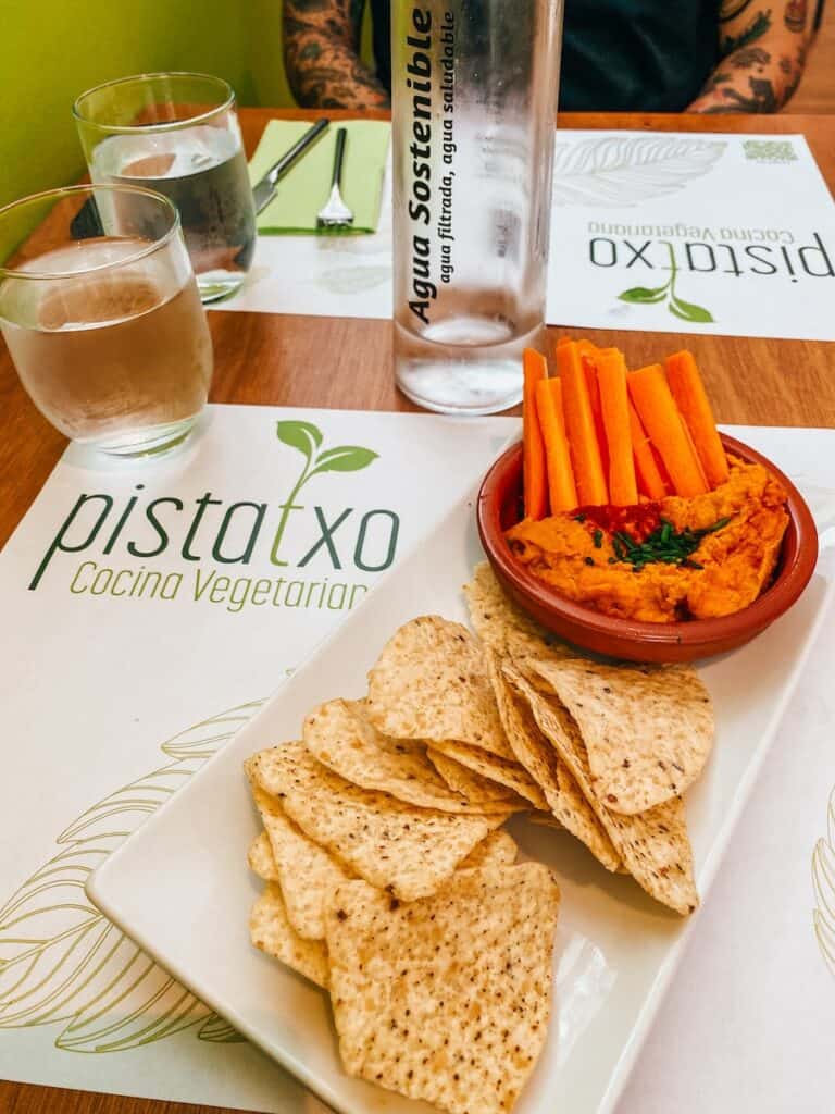 A small bowl of hummus, carrot sticks and some tortillas with a glass of water next to it