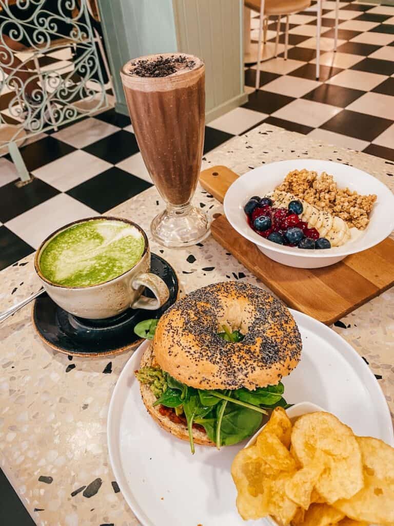 A table with different things on it, a matcha latte, a chocolate milkshake, a bowl of joghurt with fruits, and a bagel with chips next to it
