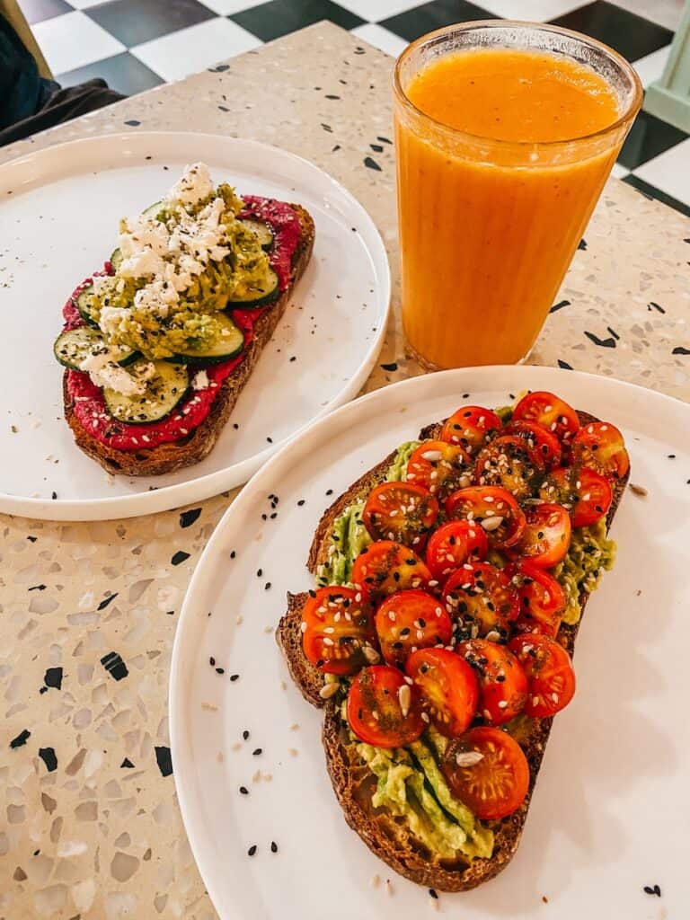 Two slices of bread, one with avocado and tomatoes on top and the other one with beetroot hummus and cucumber, next to it is a glass of orange juice
