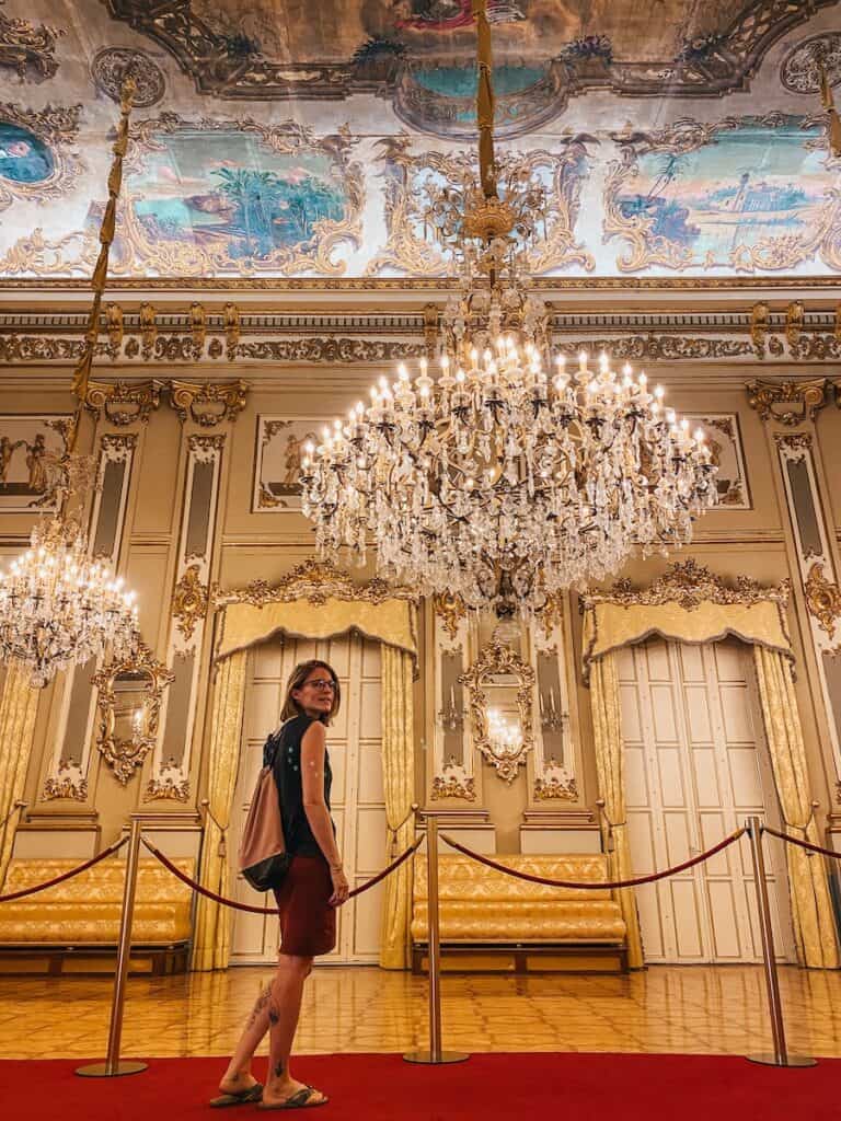 Tina standing in a very opulent room with a big chandelier and richly decorated walls and ceiling