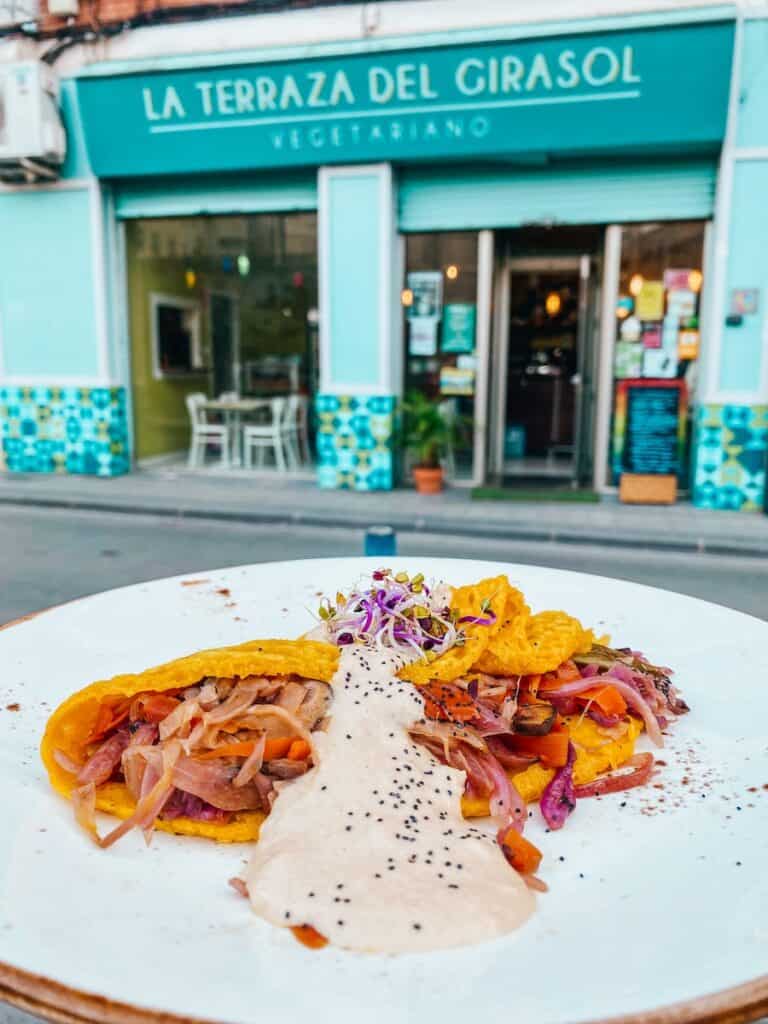 A vibrant vegan taco filled with sautéed vegetables, topped with a creamy white sauce and garnished with sprouts, served on a white plate in front of La Terraza del Girasol, a vegetarian restaurant in Murcia, Spain.