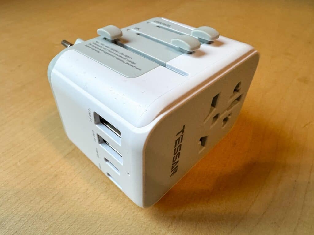 The image shows a white universal travel adapter with multiple USB ports and socket types, branded "TESSAN." It includes sliders at the top for adjusting plug types to fit various international outlets, making it suitable for different regions. The adapter sits on a light wooden surface.