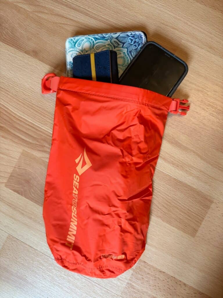 The image shows a red Sea to Summit dry bag with the brand's logo printed on it, partially open and containing a few personal items, including a smartphone, a notebook, and a tablet with a decorative case. The dry bag is placed on a light wood floor, and its lightweight and compact design is emphasized by the “Lightweight 1.5L” label near the bottom.