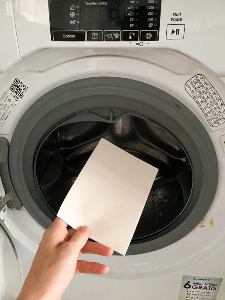 A hand holding a laundry sheet in front of the open drum of a washing machine. The machine has a digital control panel displaying various washing options and symbols, and a sticker indicating a 6-month warranty in the bottom right corner.
