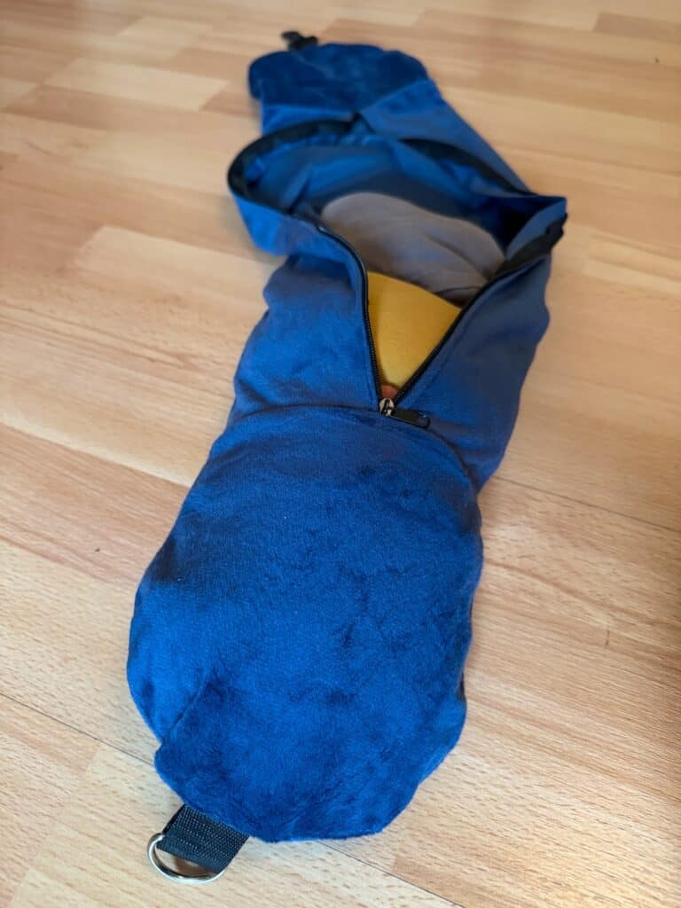 A blue travel neck pillow packed inside its travel pouch on a wooden floor. The pouch is zippered and partially open, revealing the yellow fabric of the pillow inside.