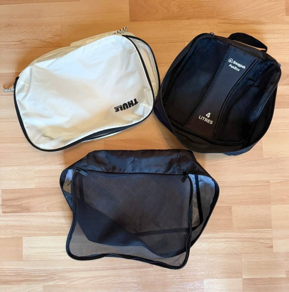 Three packing cubes of different sizes and brands placed on a wooden floor. The cubes include a white Thule cube, a black Snugpak 4-liter cube with mesh, and a larger black mesh cube.