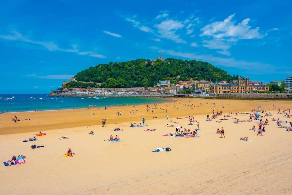 A view of the historic promenade at La Concha Beach, with elegant buildings, white balustrades, and Monte Urgull in the background. The sandy beach reflects the clear blue sky and waves of the Atlantic Ocean.