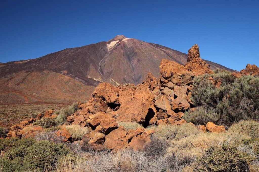 Rugged volcanic terrain with red rock formations in the foreground and the towering peak of Mount Teide in the background. The clear blue sky accentuates the dramatic landscape of this Canary Islands landmark.