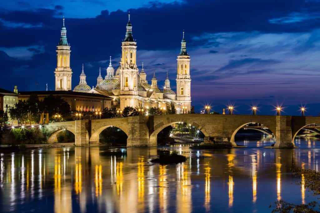 This image features the Basilica of Our Lady of the Pillar in Zaragoza, Spain, illuminated beautifully at night. The reflection of the basilica and the historic stone bridge shimmers in the Ebro River, set against a twilight sky. The scene captures the city's architectural splendor and serene ambiance.
