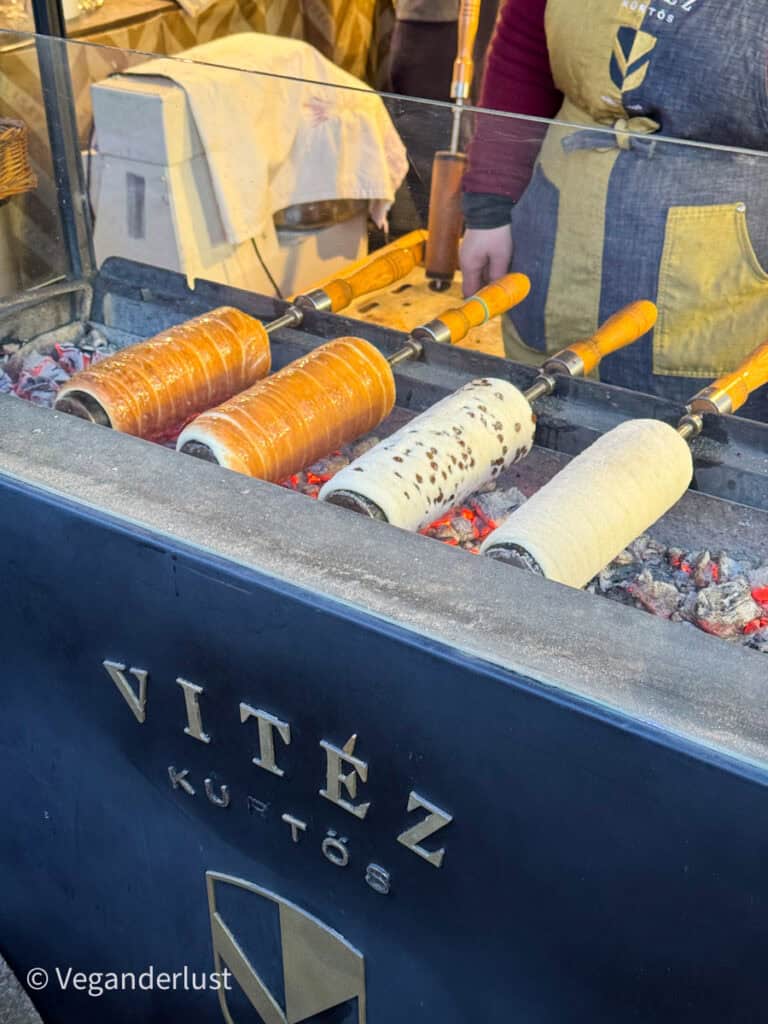 Close-up of traditional Hungarian chimney cakes (kürtőskalács) being roasted over hot coals at a market stall in Budapest. The Vitéz Kürtős branding is visible on the counter, and the cakes are turning golden brown, with some topped with sugar or chocolate.