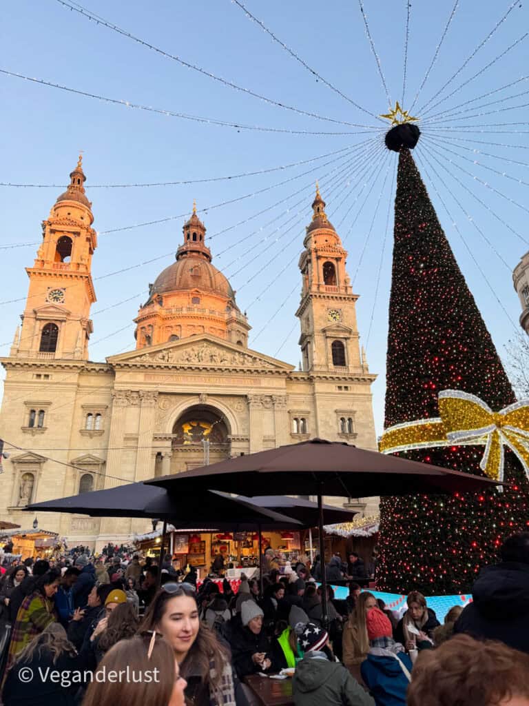 Festive Christmas market scene in front of St. Stephen's Basilica in Budapest, featuring a towering Christmas tree adorned with lights and a golden ribbon, market stalls, and a bustling crowd enjoying the holiday atmosphere under a clear evening sky.