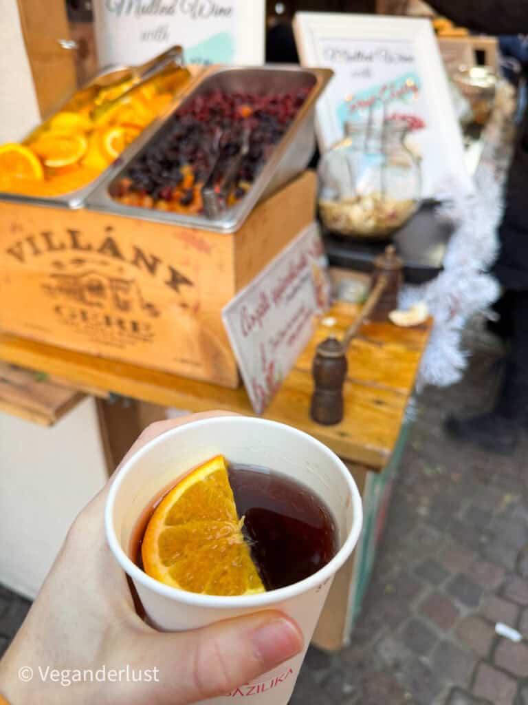 A hand holds a paper cup of mulled wine with a slice of orange, against the backdrop of a festive stand offering garnishes like oranges and dried fruits. Signs in the background promote mulled wine with local flavors at Budapest's Christmas market.