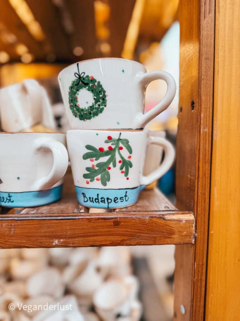 Close-up of handcrafted ceramic mugs on a wooden shelf, painted with festive designs like a wreath and mistletoe, and the word 'Budapest' written at the bottom. The background features warm lighting from a Christmas market stall.