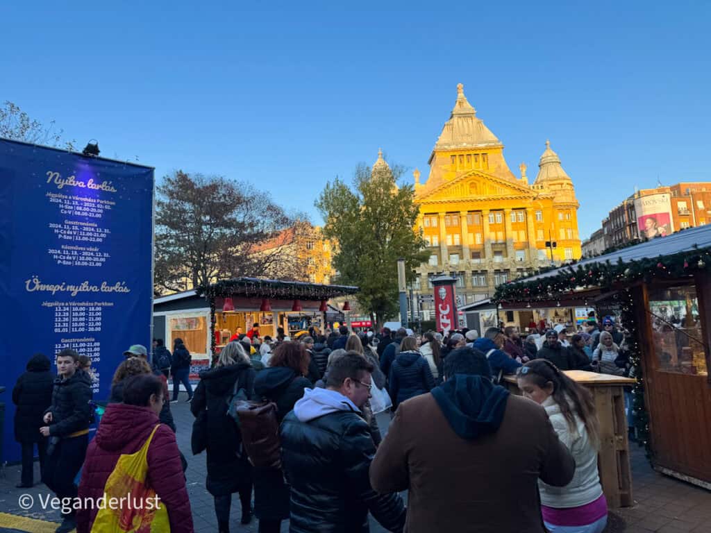 Crowd gathered at Budapest's Vörösmarty Square Christmas market, with festive stalls and a large informational sign displaying market hours in Hungarian. The illuminated yellow facade of a historic building contrasts with the clear blue sky in the background.