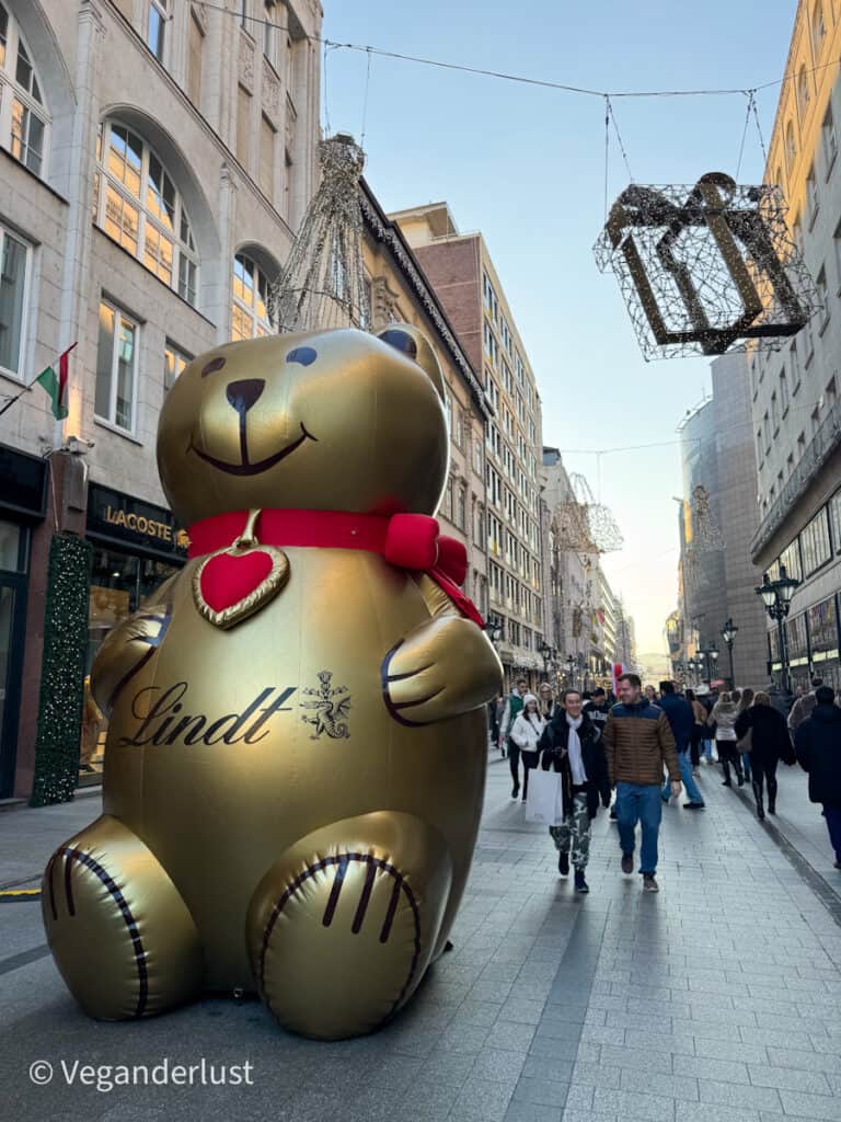 A giant golden inflatable Lindt bear with a red bow and heart decorates Deák Ferenc Street in Budapest, surrounded by festive hanging lights shaped like gifts. Shoppers and pedestrians stroll along the vibrant holiday-themed street.
