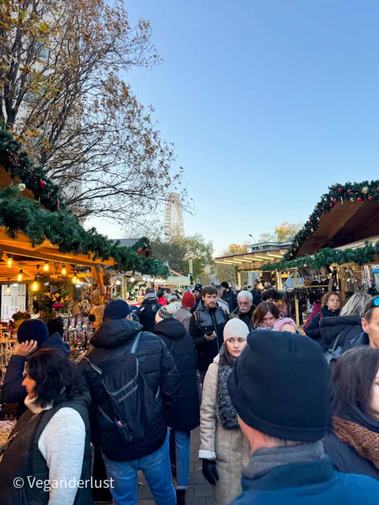 A bustling scene at Budapest's Erzsébet Square Christmas market, with crowds browsing festive stalls adorned with garlands. The Budapest Eye Ferris wheel rises in the background against a clear blue sky.