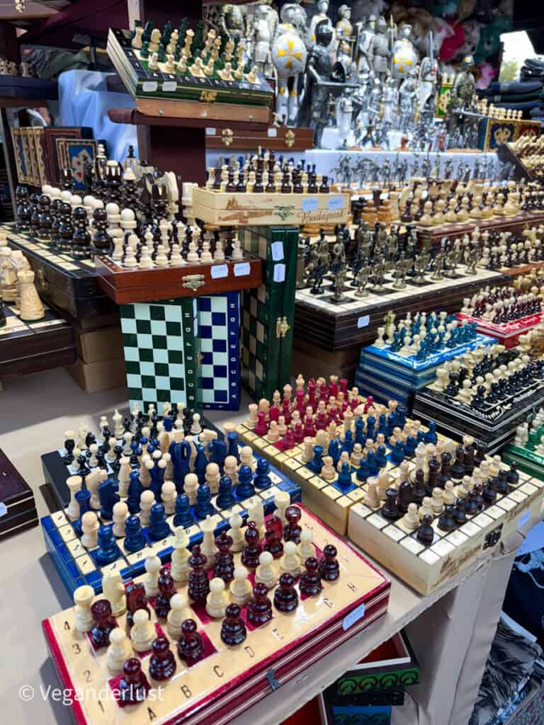 A market stall at Budapest's Christmas market displaying an array of chess sets in different colors and styles, with some boards featuring Budapest-themed designs. The background includes silver knight figurines and other handcrafted ornaments.