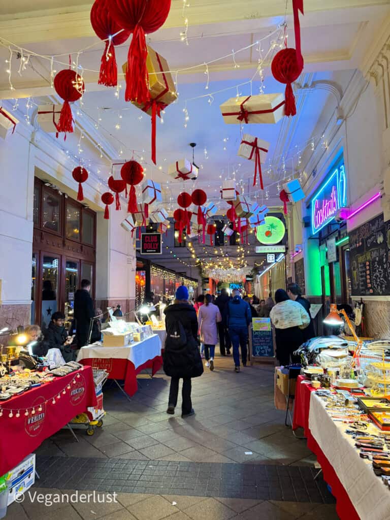 A brightly lit corridor at Gozsdu Court Christmas market in Budapest, decorated with hanging red lanterns, wrapped gift boxes, and twinkling fairy lights. Visitors stroll through while exploring stalls selling festive gifts and crafts.