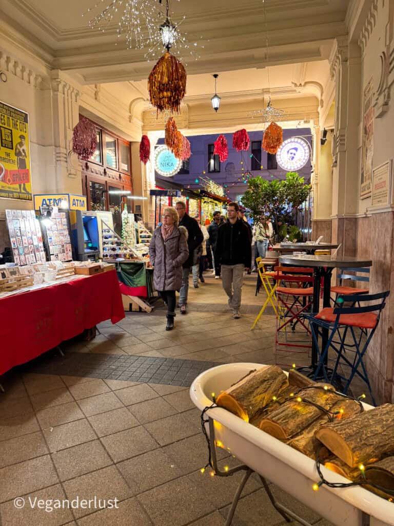 A cozy market corridor at Gozsdu Court in Budapest, adorned with sparkling string lights and red metallic tinsel decorations. Market stalls line the walkway, showcasing crafts and souvenirs, while visitors browse under the festive lighting.