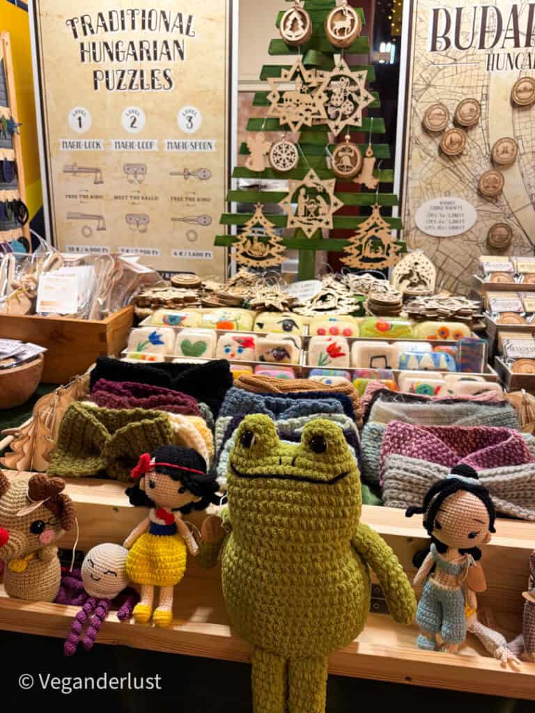 A market stall displaying handmade items at Gozsdu Court Christmas market, featuring crocheted toys like a green frog and Snow White, colorful scarves, and wooden Hungarian puzzles. Wooden ornaments shaped like stars and trees are displayed on a green stand nearby.