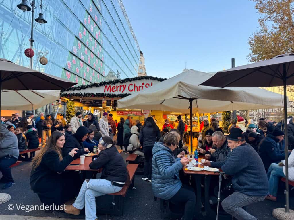 Crowds enjoy food and drinks at outdoor tables under umbrellas at the Vörösmarty Square Christmas market in Budapest, with a 'Merry Christmas' sign above a festive food stall and reflections of Christmas-themed decorations on a glass building in the background.