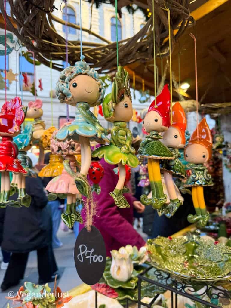 Colorful handmade ceramic ornaments featuring whimsical fairies and gnomes hang at a Budapest Christmas market stall, with a price tag showing '8700' in Hungarian Forints. A wicker wreath decorates the stall, and people browse in the background.