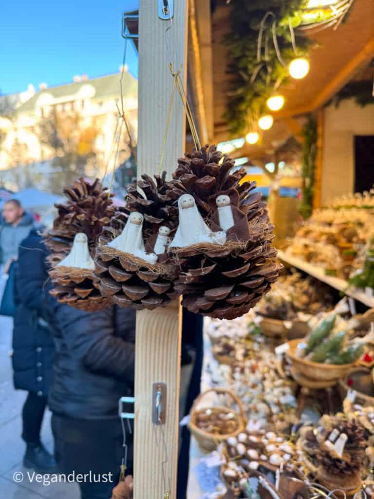 Handmade Christmas ornaments featuring tiny nativity scenes crafted from pinecones, with miniature figures representing Mary, Joseph, and baby Jesus. The ornaments hang on a wooden display at a festive market stall in Budapest, surrounded by baskets of natural and rustic holiday decorations.