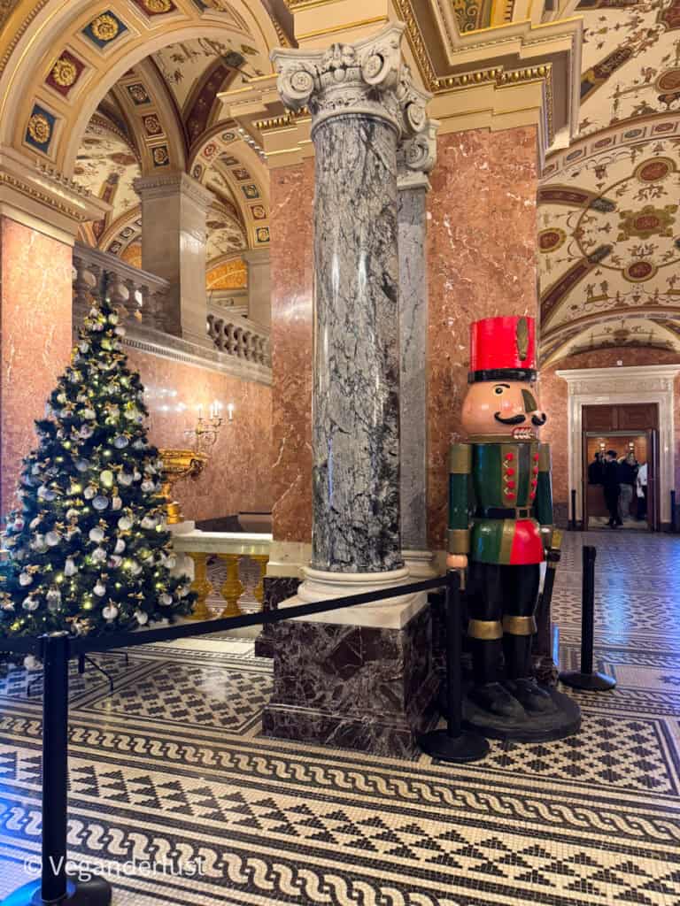 The opulent interior of the Hungarian State Opera House in Budapest, showcasing a grand marble column, ornate ceilings, and a decorated Christmas tree with silver ornaments. A life-sized wooden Nutcracker statue stands nearby on the patterned floor.