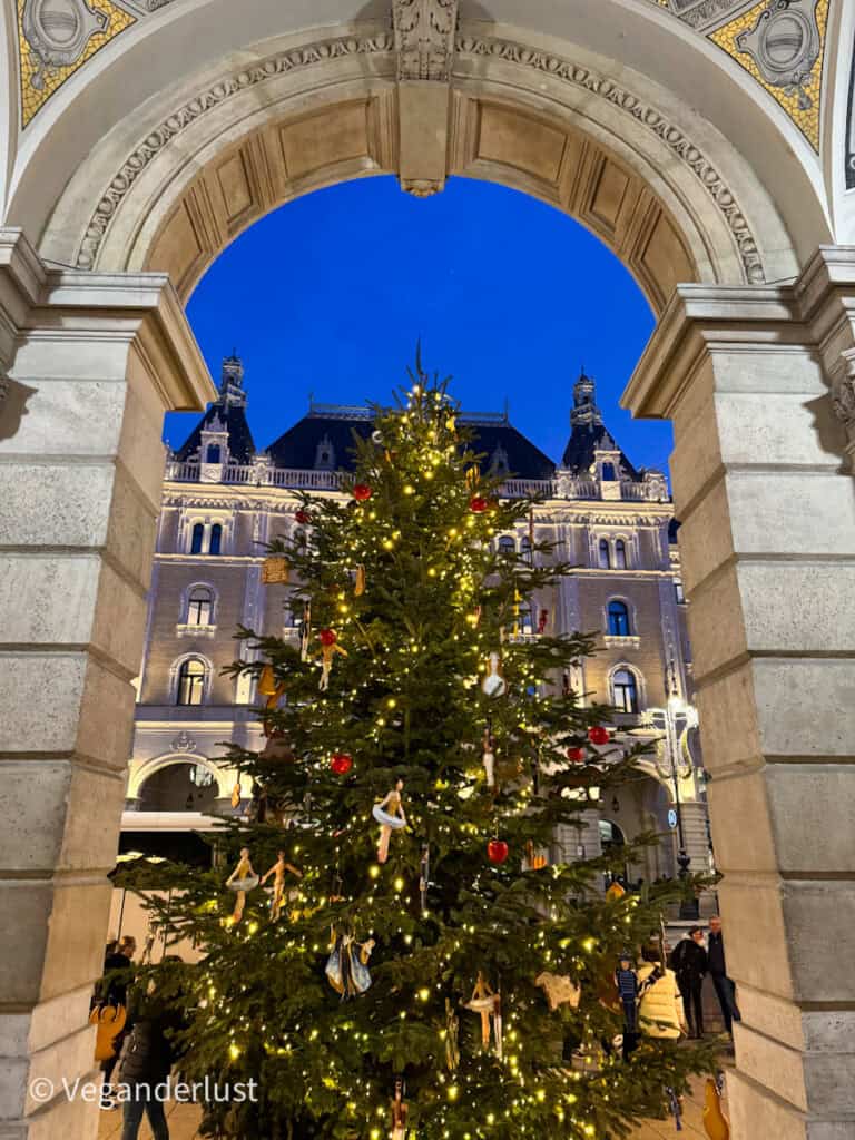 A large, beautifully decorated Christmas tree adorned with lights and ornaments, framed by an ornate archway of the Hungarian State Opera House in Budapest. The illuminated facade of a historic building provides a striking backdrop against the deep blue evening sky.