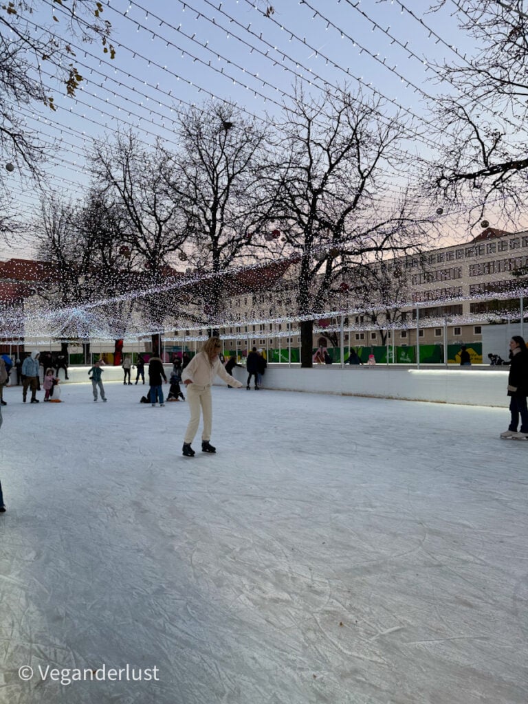 Ice skating rink in Budapest surrounded by bare winter trees decorated with twinkling string lights, creating a festive canopy. Skaters of all ages enjoy the rink under a soft evening sky, with nearby buildings visible in the background.