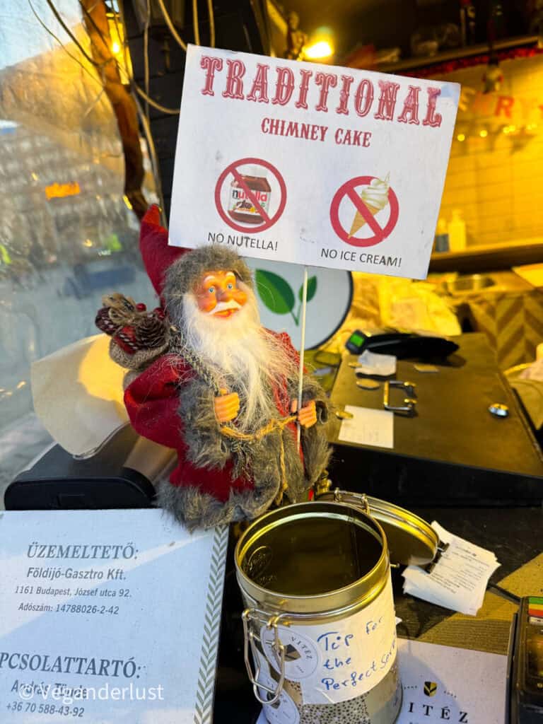 Close-up of a festive stall at a Budapest market, featuring a small Santa Claus figure holding a sign that reads 'Traditional Chimney Cake' with icons indicating 'No Nutella!' and 'No Ice Cream!' A nearby tip jar labeled 'Tip for the perfect service' sits on the counter under warm lighting.
