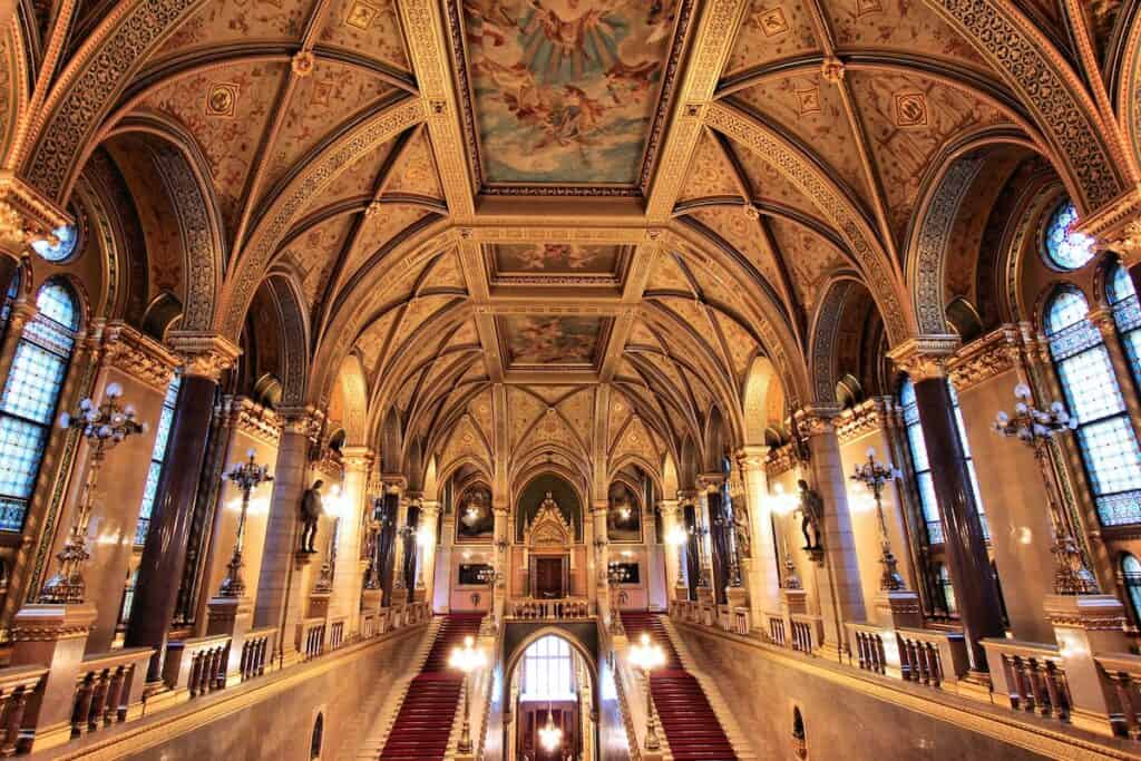 The grand interior of Budapest's Parliament building, featuring a stunning vaulted ceiling adorned with intricate patterns and frescoes. The hall is illuminated by ornate chandeliers and lined with tall stained-glass windows, red-carpeted staircases, and statues, showcasing the building's opulent architectural design.