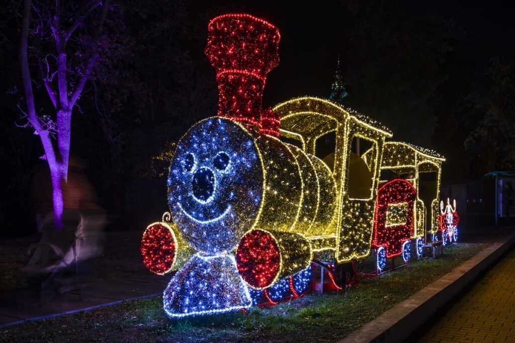 A festive display of a train made entirely of colorful Christmas lights, including a smiling face on the engine, set against a dark park background with faint silhouettes of trees.