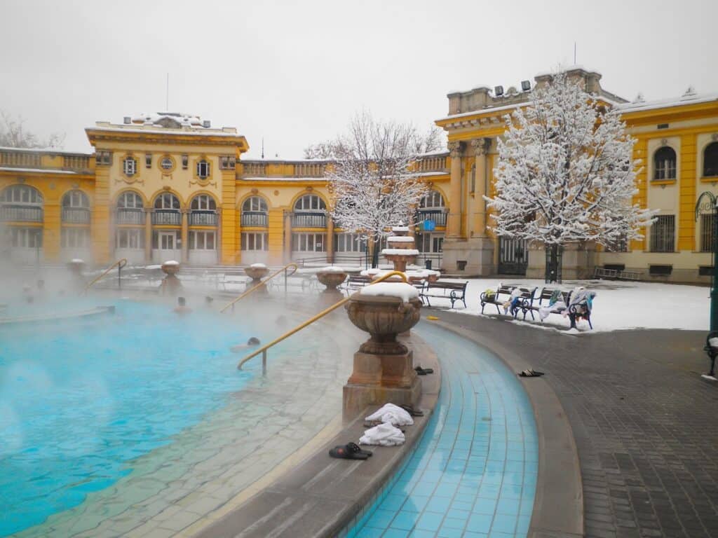 The outdoor thermal pool at Budapest's Széchenyi Baths during winter, with steam rising from the warm water as swimmers relax. The surrounding yellow Neo-Baroque building and snow-covered trees create a picturesque and serene winter scene.