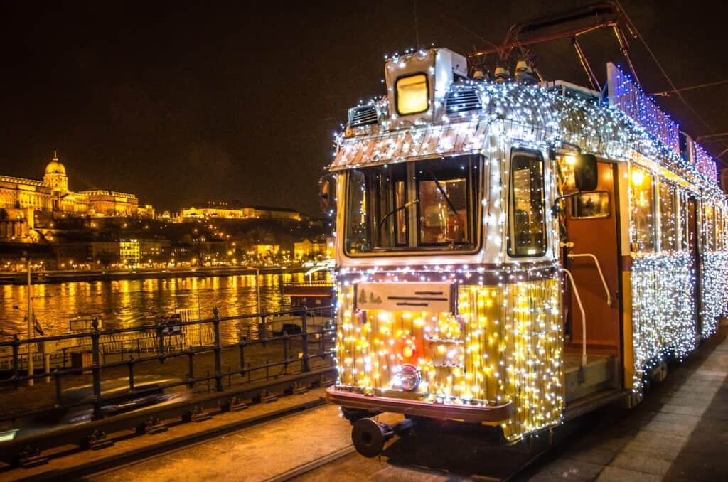 A tram in Budapest covered in glowing white Christmas lights, parked by the Danube River with the illuminated Buda Castle in the background reflecting off the water.