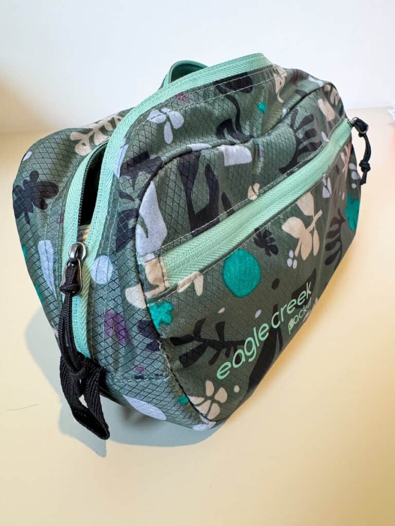 A green floral-patterned Eagle Creek toiletry bag with mint green zippers sits on a light-colored surface. The bag has a slightly bulging shape, suggesting it is filled with items. The "Eagle Creek Pack-It" logo is visible on the front pocket.