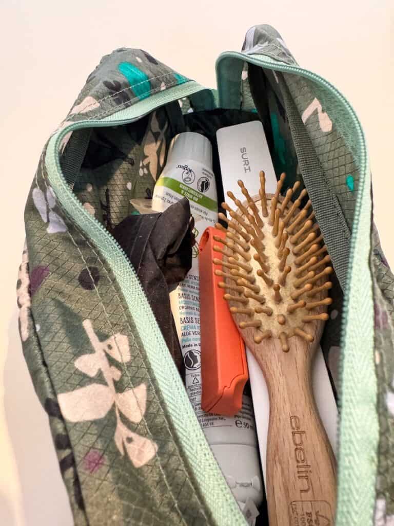The same Eagle Creek toiletry bag is open, revealing various travel essentials inside. Items include a wooden hairbrush labeled "ebelin," a white rectangular case marked "SURI," a small bottle of deodorant, a tube of cream, and other toiletries. The bag’s patterned fabric and mint green zipper contrast with the contents inside.