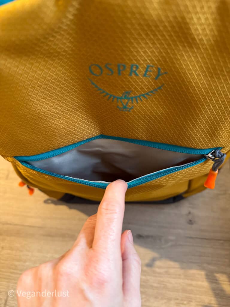 Close-up of a yellow Osprey Daylite backpack with teal zippers, showing the small front pocket partially unzipped while a hand gently holds it open, highlighting the smooth fabric interior.